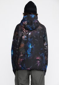 Veste à motif floral noir avec des couleurs vives. Dotée d'une capuche et d'un tissu texturé. Portée avec un pantalon gris, offrant une coupe décontractée.