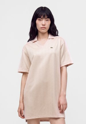 Giovane donna con lunghi capelli neri che indossa un vestito polo beige chiaro a maniche corte con un piccolo logo verde sul petto.