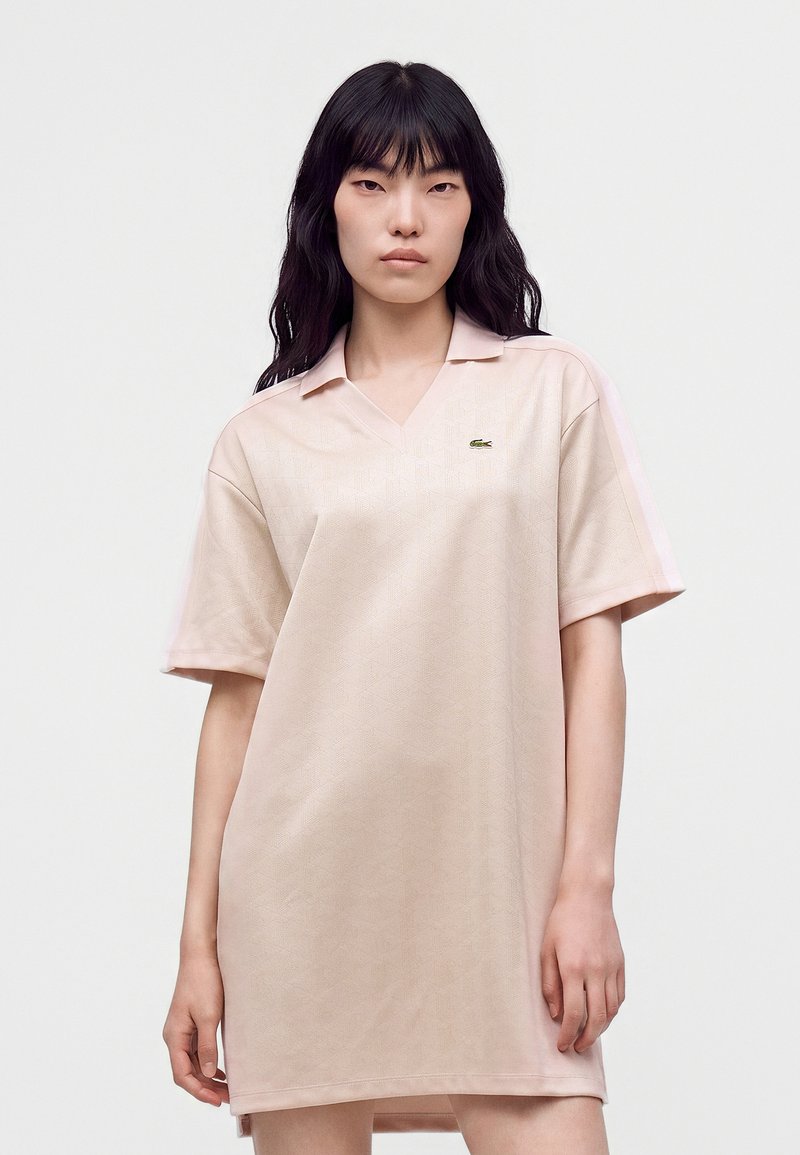 Jeune femme aux longs cheveux noirs portant une robe polo beige clair à manches courtes avec un petit logo vert sur la poitrine.