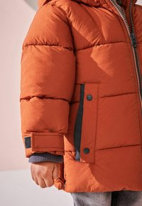 Veste orange matelassée avec une fermeture éclair, des boutons noirs en accent, et un tissu texturé. Comprend une capuche et des poignets côtelés pour plus de chaleur.