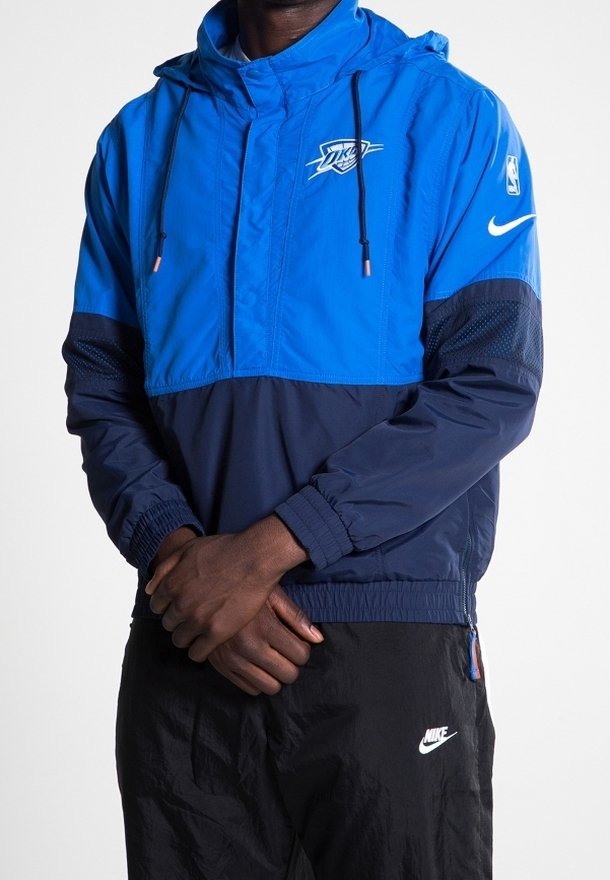 Homme portant un coupe-vent bleu et marine Oklahoma City Thunder Nike et un pantalon noir Nike, debout les mains jointes devant.