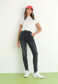 Jeans skinny de tiro alto en negro con detalles de tachuelas plateadas, combinados con una camiseta blanca y una gorra naranja, llevados con zapatillas blancas.