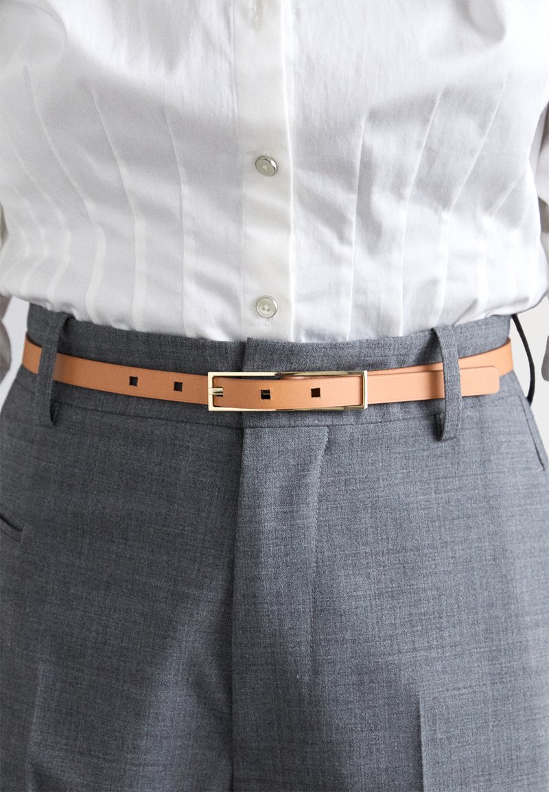 Brūns ādas josta ar taisnstūra zelta sprādzi ar kvadrātveida izgriezumiem, nēsāta pār pelēkiem teksturētiem biksēm un baltu krekla ar apkakli.