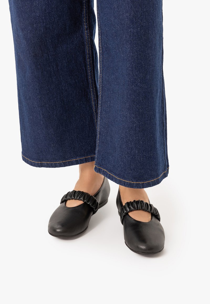 Pieds portant des ballerines en cuir noir avec des brides froncées, associées à un jean denim bleu foncé à jambes larges sur fond blanc.