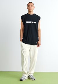 Črna brezrokavna majica Nike Air z belim logotipom; kombinirana z ohlapnimi belimi hlačami in sivimi supergami. Gladka tekstura, sproščen oblikovni slog.