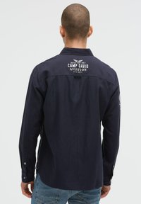 Marineblaue Langarmbluse mit spitzem Kragen, versehen mit einem Logo auf der Rückseite und Knopfdetails an den Manschetten. Gewebte Struktur.