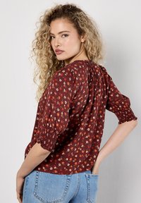Blusa rossa floreale con maniche a sbuffo corte, schiena arricciata e vestibilità ampia. Presenta un motivo di piccoli fiori su un tessuto texturizzato.