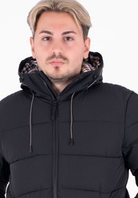 Giacca a collo alto nera in stile puffer con cappuccio, dotata di chiusura a zip e lacci regolabili. Fodera interna a quadri visibile attorno al collo e al viso.