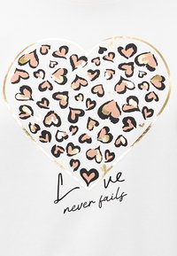 Witte graphic tee met een groot hartontwerp gevuld met roze, zwarte en gouden luipaardprints. De tekst luidt "Liefde faalt nooit" in het zwart.