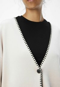 Lyse beige cardigan med sort syning langs v-halsen, med en sort knap. Båret over en struktureret sort ribbet top.
