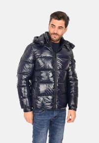Giubbotto puffer blu navy con finitura lucida, motivo a cassettoncini orizzontali, cappuccio attaccato, zip frontale e tasche laterali. Indossato sopra un maglione scuro.