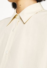 Chemise beige clair à boutons avec col classique. Fabriquée en tissu lisse, elle présente une patte de boutonnage dissimulée et un léger éclat.