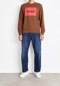 Sweat-shirt marron avec un logo rectangulaire rouge affichant « HUGO » en noir, assorti à un jean en denim bleu et des baskets blanches. Coupe décontractée.