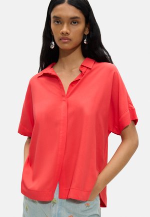 Młoda kobieta z długimi czarnymi włosami, ubrana w czerwoną koszulę z krótkim rękawem i guziczkami oraz jasnoniebieskie spodnie z kwiatowym haftem, ręce w kieszeniach.