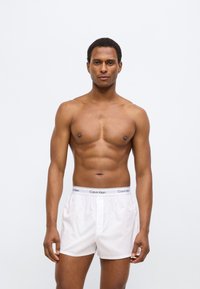 Άσπρο μπόξερ με ελαστική μέση Calvin Klein, λείο ύφασμα και άνετη εφαρμογή, φορεμένο από άνδρα μοντέλο με αθλητική σωματική διάπλαση.