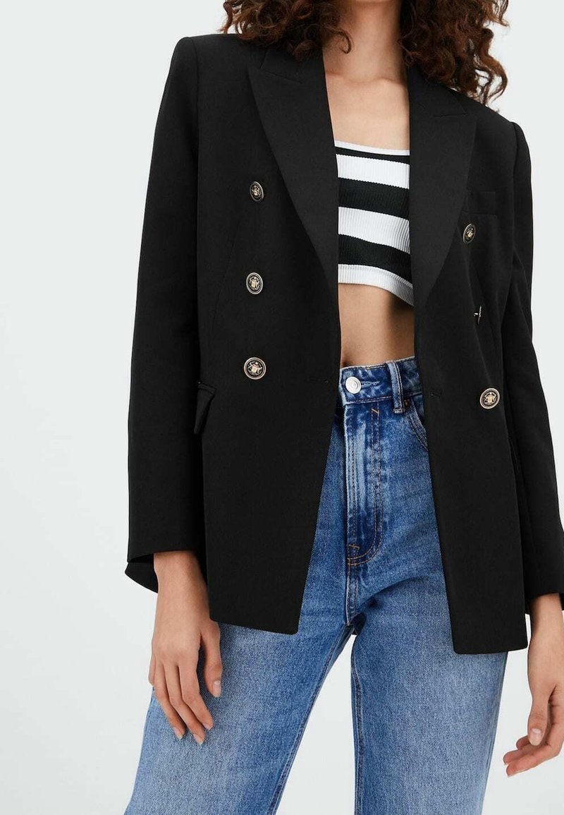 Femme portant un blazer noir croisé à boutons dorés, un crop top rayé noir et blanc, et un jean bleu taille haute.