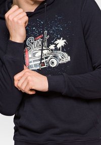 Černá mikina s kapucí s grafikou starožitného auta, palmami a nápisem "Summer Surf". Materiál vypadá měkce s hladkou texturou.