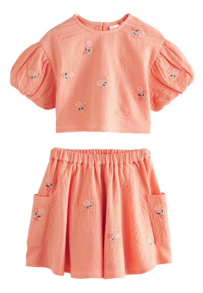 Korálový dvoudílný outfit s nafouknutými rukávy; vyšívané květinové detaily na topu a sukni. Sukně má elastický pas a boční kapsu.