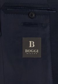 Intérieur de blazer bleu foncé montrant les détails du bouton, de la poche, et une étiquette noire avec "B BOGGI MILANO" brodée à l'intérieur.