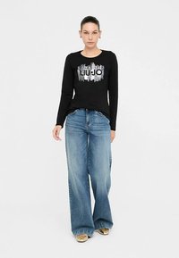 Črna majica z dolgimi rokavi z grafičnim dizajnom, kombinirana s svetlo modrimi širokimi kavbojkami in zlatimi supergami. Enostaven in priložnosten videz.