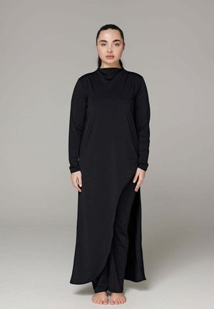 Femme debout pieds nus portant une longue robe noire à col montant, manches longues et fente devant, par-dessus un pantalon noir.