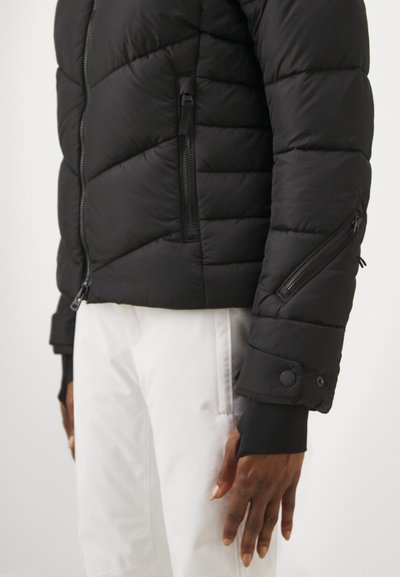 Μαύρο φουσκωτό μπουφάν με καπιτονέ σχέδιο, διαθέτει τσέπες με φερμουάρ και μανίκια με κουμπί, συνδυασμένο με λευκό παντελόνι.