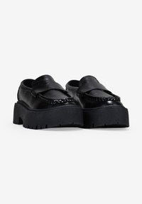 Mocasines de plataforma de cuero negro con un upper texturizado, diseño sin cordones y suela gruesa estriada. Presenta costuras decorativas en la parte frontal.