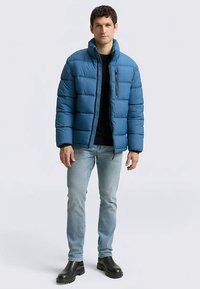 Blauer Steppdaunenmantel mit Steppmuster, hohem Kragen und links einer Reißverschlusstasche. Kombiniert mit hellen Jeans und schwarzen Stiefeln.