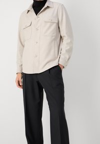 Chaqueta beige de botón con dos bolsillos en el pecho, hecha de un material suave. Combinada con pantalones rectos negros, que presentan una textura suave.