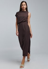 Robe marron côtelée sans manches avec une taille froncée et une fente latérale. Associée à des sandales légères tissées et des boucles d'oreilles simples. Texture lisse.