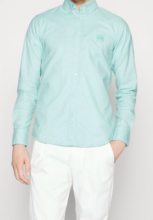 Mann trägt ein hellblaues, langärmeliges Hemd mit Knöpfen und einem kleinen Logo auf der Brust sowie weiße Hose, eine Hand in der Tasche.