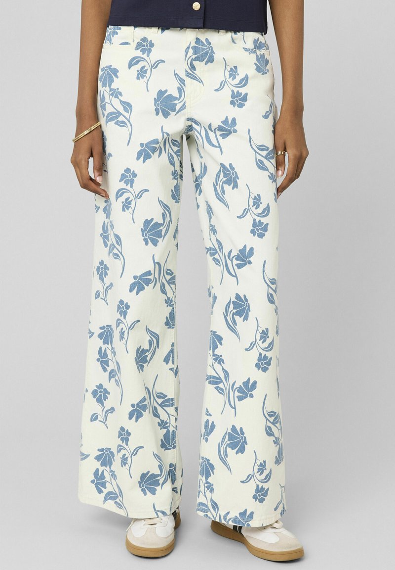Femme portant un pantalon blanc à jambes larges avec un imprimé floral bleu, des baskets blanches, un bracelet en or et un haut bleu marine.