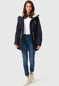 Marinefarbener Parka mit einer weißen Fleece-gepolsterten Kapuze, verstellbaren Kordelzügen und braunen Akzenten; kombiniert mit blauen Jeans und weißen Turnschuhen.
