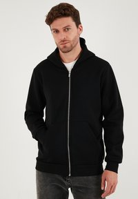 Černá mikina s kapucí na zip, prostornou kapucí, předními kapsami, žebrovanými manžetami a měkkou texturou látky. Model ji nosí spolu se šedými džíny.