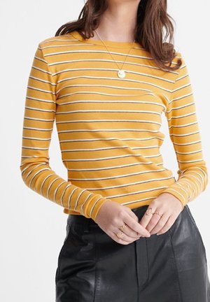 Woman wearing mustard yellow long-sleeve shirt with white stripes and black leather pants, accessorized with gold rings and a pendant necklace.