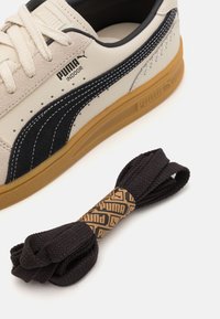 Puma Hallenschuhe aus cremefarbenem Leder mit schwarzen Wildlederakzenten, ausgestattet mit einer Gummisohle. Ein schwarzes Schuhband und eine gebrandete Verpackung sind enthalten.