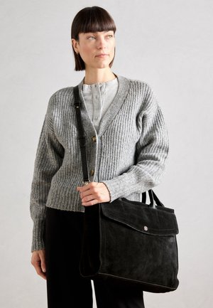 Kvinde med kort brunt hår iført en grå strikcardigan og holder en stor sort ruskind skuldertaske.
