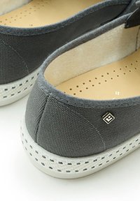Chaussons en tissu gris à bout rond, avec une semelle en caoutchouc blanche et une semelle intérieure perforée. Présente un petit logo métallique sur le côté.
