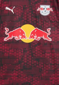 Rød og sort sportsjersey med Puma-logo, RB Leipzig-skilt og to røde biler, der vender mod en gul cirkel, med et geometrisk mønster i baggrunden.