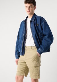 Denim bomber jacket in dark blue with elastic cuffs; white t-shirt underneath; beige cargo shorts with side pockets and button closure.