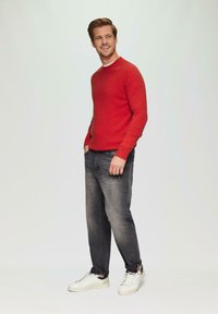 Rode ronde hals trui, grijze jeans en witte sneakers. De trui heeft ribbelmanchetten; de jeans hebben een vervaagde afwerking. Eenvoudige, casual outfit.