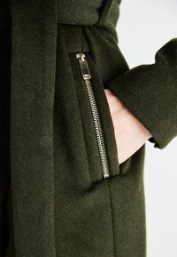 Cappotto verde in lana con una tasca con zip in tonalità dorata, texture morbida e dettaglio con cintura in vita.