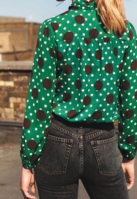 Grøn bluse med store brune og hvide prikker, med høj krave og pufærmer, kombineret med høj talje mørke denimjeans.
