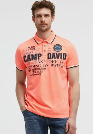Mann trägt ein helloranges Polohemd mit dunkelblauem Text und Grafiken, kombiniert mit blauer Jeans, steht mit einer Hand in der Hosentasche.
