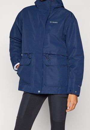 Persona que lleva una chaqueta con capucha azul de Columbia con grandes bolsillos delanteros y mallas negras, de pie con una mano en el bolsillo.