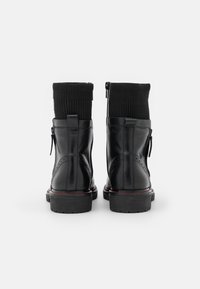 Schwarze Lederstiefeletten mit gerippten, schwarzen, sockenähnlichen Schaft, seitlichen Reißverschlüssen und roten Nähten auf dicken, schwarzen Sohlen. Glatte Textur, abgerundete Form.