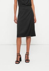 Schwarzer knielanger Rock mit subtiler Textur und kleinen Punktmustern, kombiniert mit schwarzen Riemchen-High-Heelsandalen mit stabilen Absätzen.