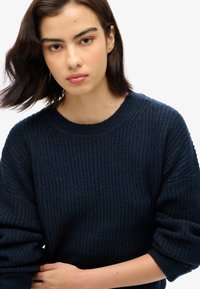 Navy textured knit sweater with a round neckline and slightly oversized sleeves, featuring ribbed cuffs and a relaxed fit.