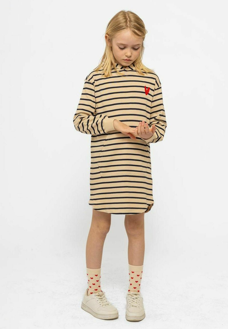 Beige randig klänning med lång ärm, prydd med ett rött hjärt detaljer på bröstet. Matchad med krämfärgade strumpor prydda med röda hjärtan och vita sneakers.
