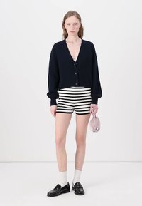 Marineblå kort cardigan med knapper, stribede shorts og sorte loafers. Model der holder en lille pink taske, med hvide sokker synlige.